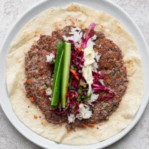A plate with a flatbread topped with ground meat, rice, red cabbage slaw, cucumber sticks, and a drizzle of white sauce.