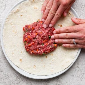 Hands pressing raw ground meat mixture with chopped vegetables onto a large tortilla on a plate.