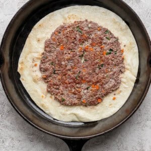 A cast iron skillet with flatbread topped with a spread of seasoned ground meat and chopped vegetables.