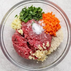A glass bowl containing ground beef, chopped parsley, diced carrots, minced garlic, breadcrumbs, and salt on a light countertop.