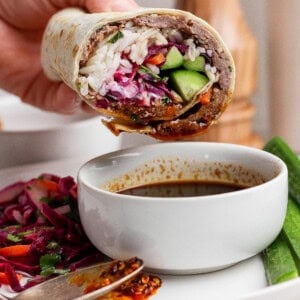 A hand holds a partially eaten wrap filled with rice, vegetables, and sauce above a white bowl of dipping sauce on a plate with salad and cucumber slices.