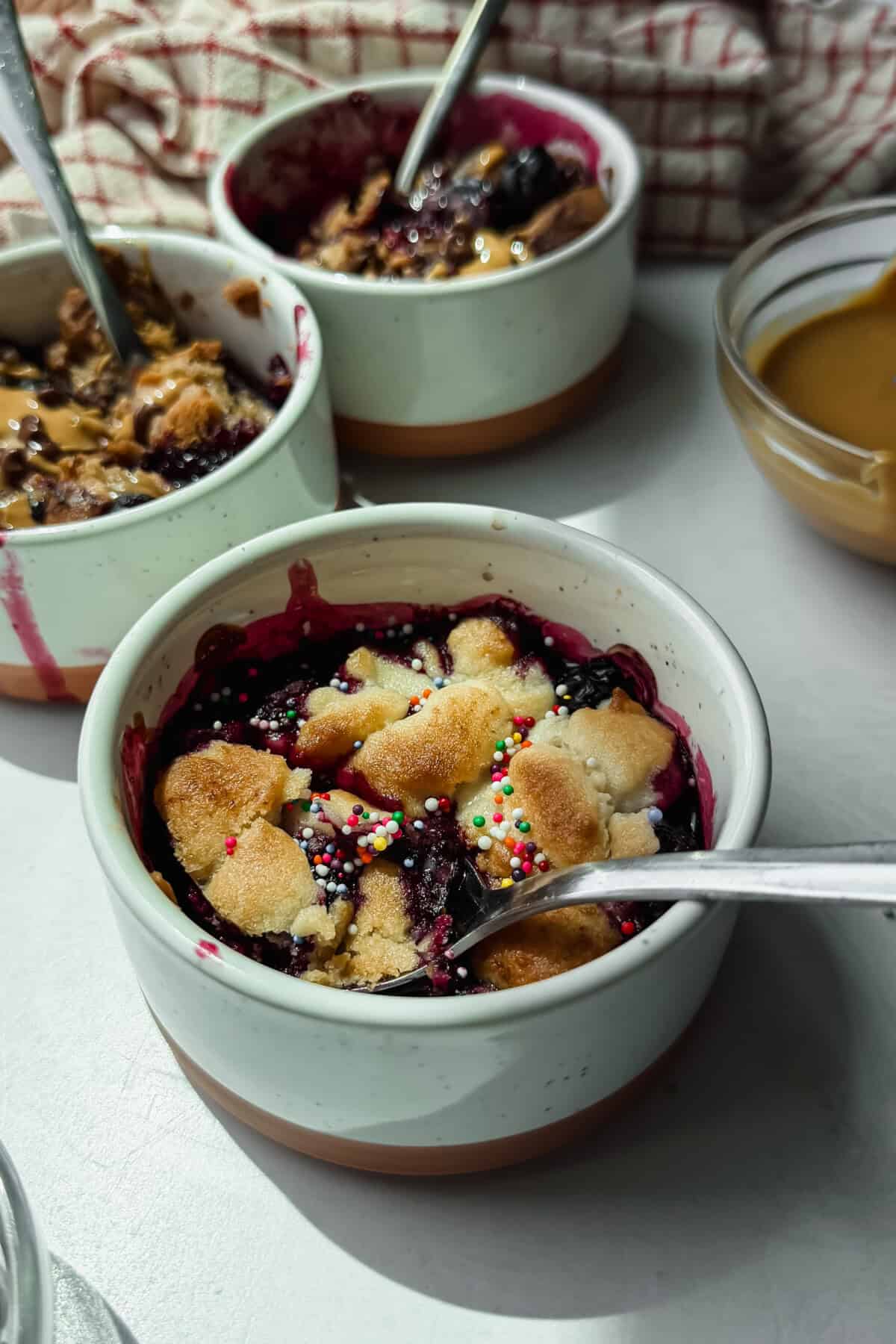 Three ceramic bowls of berry crumble topped with sprinkles and shortbread pieces, each with a spoon, sit on a light surface next to a bowl of caramel sauce.
