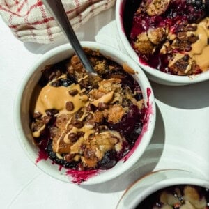 Three small white ramekins filled with baked berry dessert, topped with peanut butter and chocolate chips, with metal spoons and a red-striped cloth nearby.