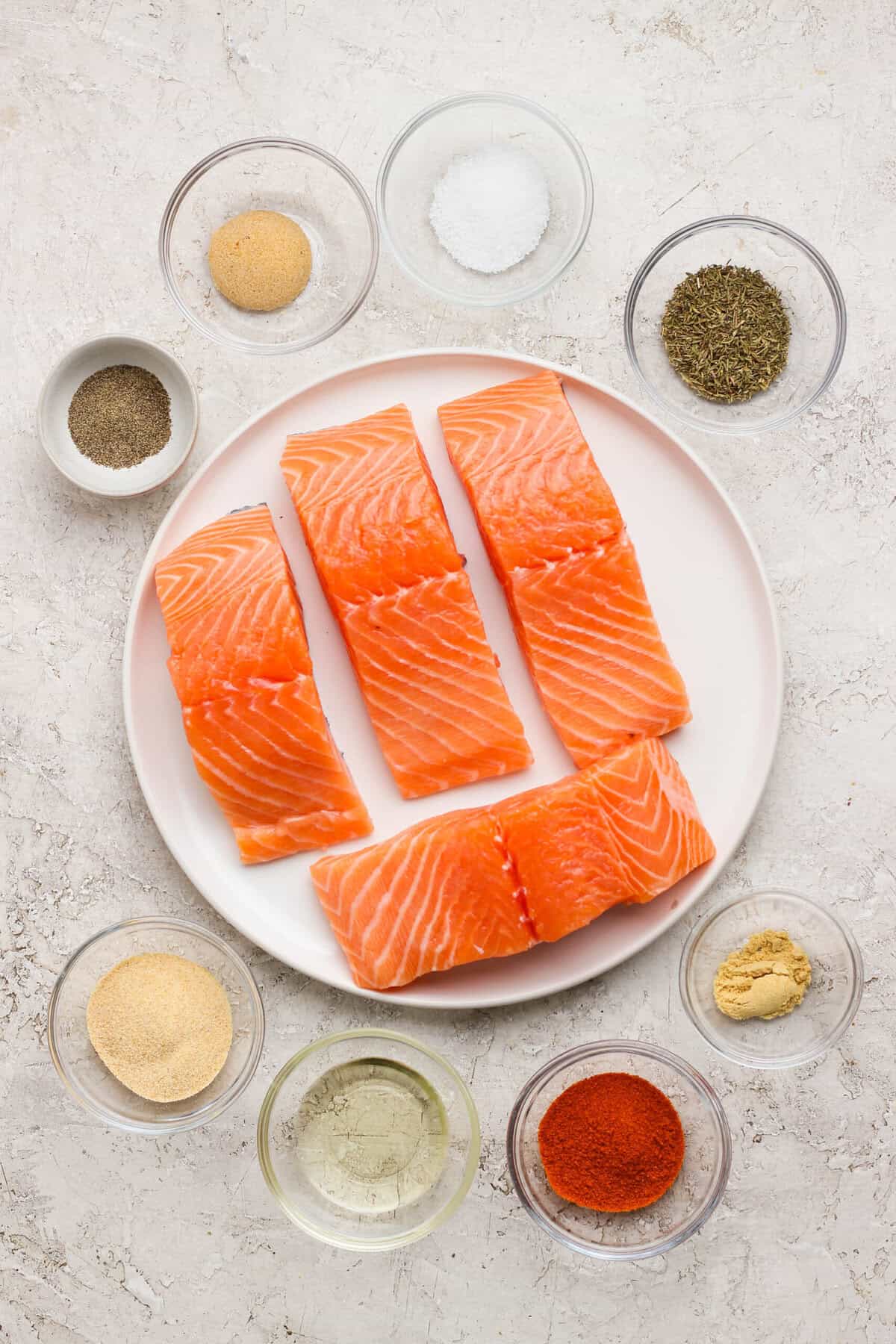 Raw salmon fillets on a plate surrounded by small bowls of seasoning, avocado oil, and herbs on a light countertop.