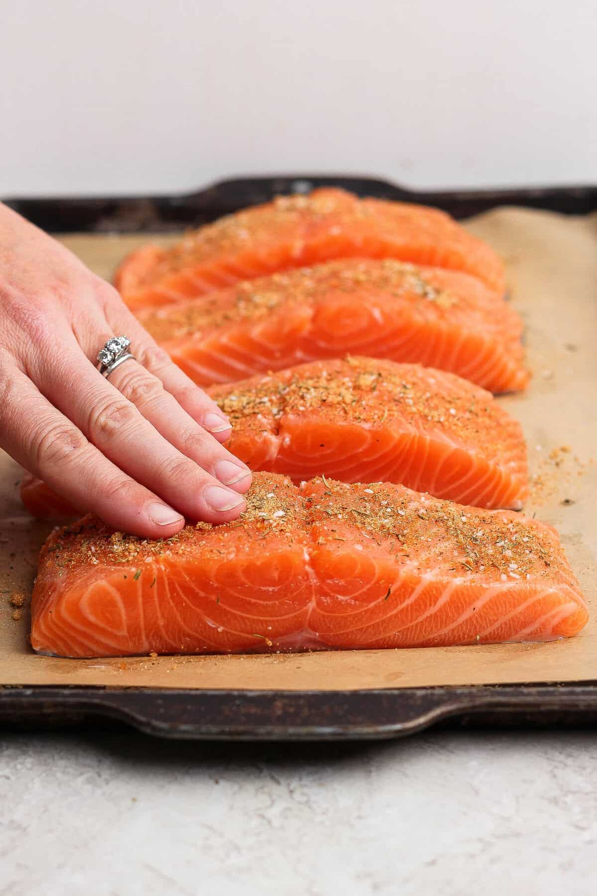 Rubbing seasoning onto raw salmon fillets on a parchment-lined baking sheet.