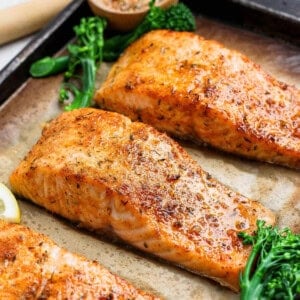 Three golden broiled salmon fillets on a parchment-lined baking sheet with broccolini and lemon.