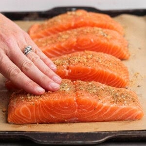 Rubbing seasoning onto raw salmon fillets on a parchment-lined baking sheet.