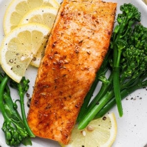 Broiled salmon fillet served on a white plate with lemon slices and steamed broccolini.