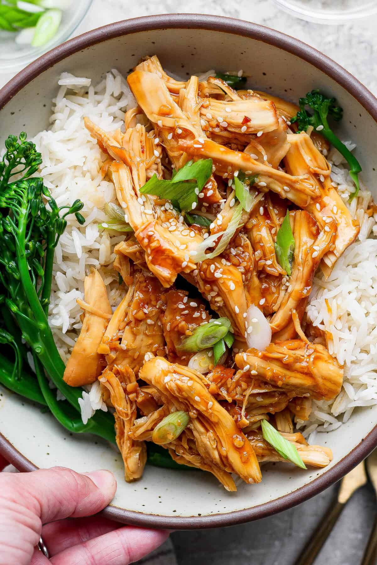 Hand holding a bowl of crockpot honey garlic chicken with rice, broccolini, and sesame seeds