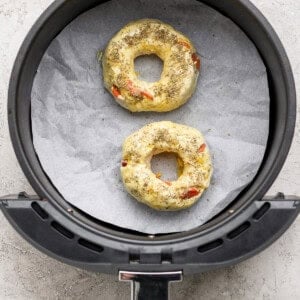 Two uncooked pizza protein bagels with seasoning in an air fryer basket