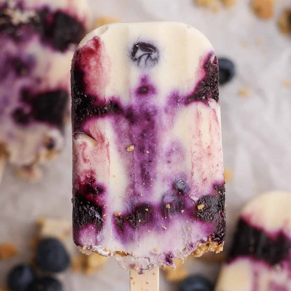 Close-up of a blueberry and cream popsicle with visible fruit swirls and a crumbly base, held upright on a wooden stick.
