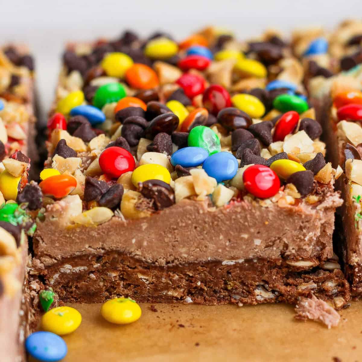 Close-up of dessert bars topped with colorful candy-coated chocolates, chocolate chips, and chopped nuts on a brown surface.