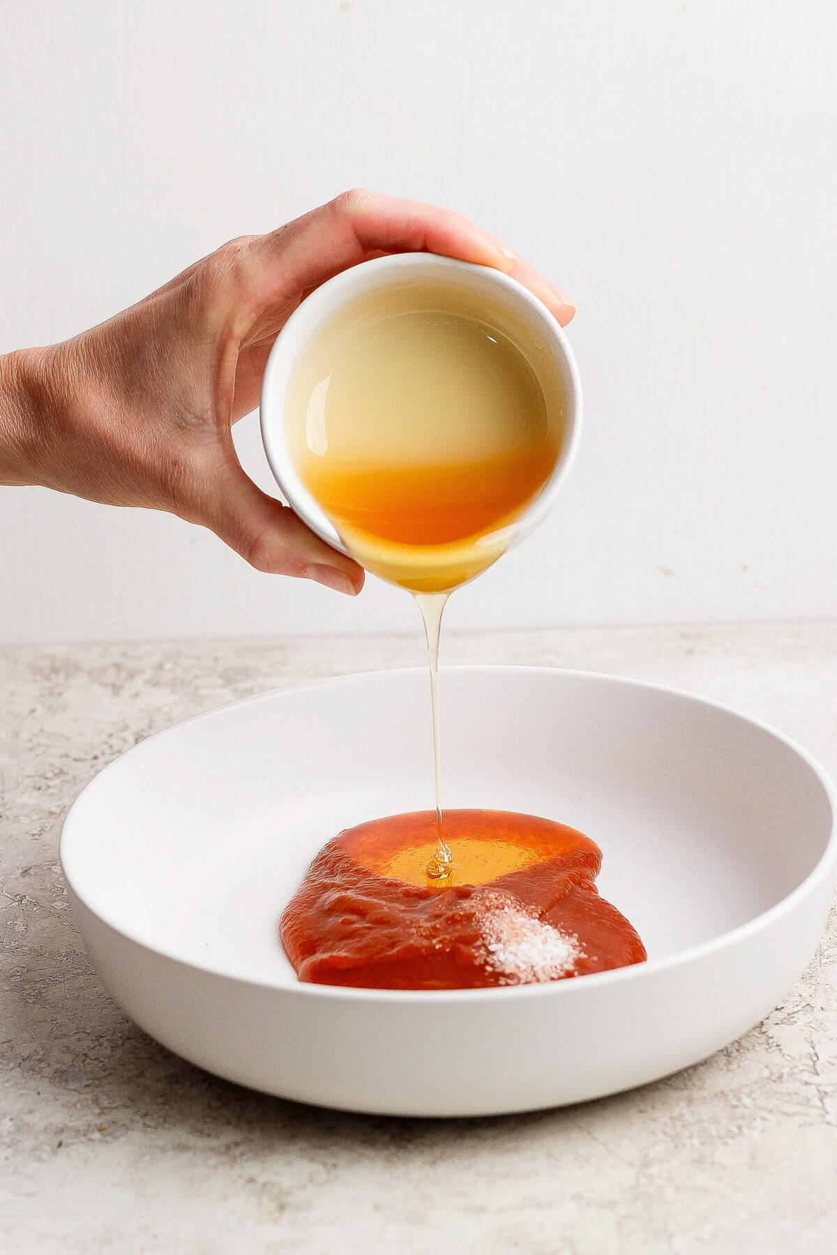 A hand pours honey from a small white bowl onto a plate of barbecue sauce.