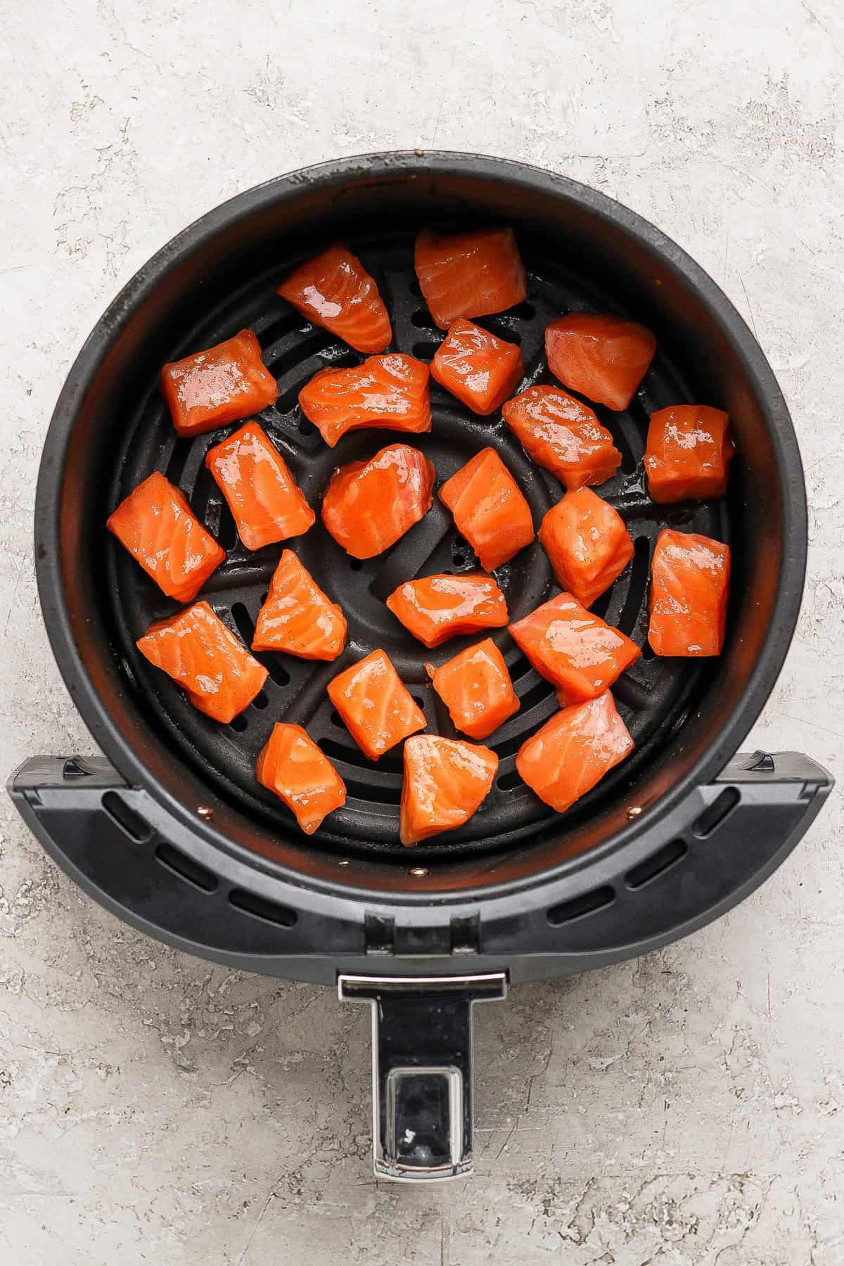 Raw salmon cubes are arranged in a single layer inside the basket of an air fryer on a light-colored surface.