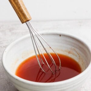 A metal whisk with a wooden handle sits in a white ceramic bowl containing a reddish-brown liquid.