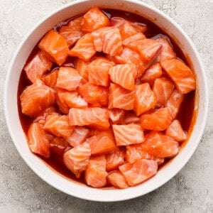 A white bowl filled with cubed raw salmon mixed with a red marinade, placed on a light textured surface.