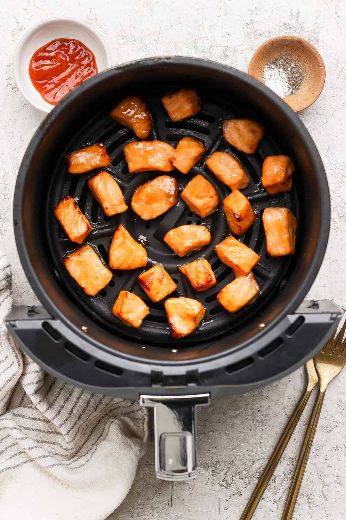 Cooked salmon pieces in an air fryer basket with bowls of sauce and seasoning nearby, a striped towel, and two forks on a light surface.