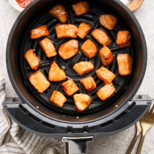 Cooked salmon pieces in an air fryer basket with bowls of sauce and seasoning nearby, a striped towel, and two forks on a light surface.