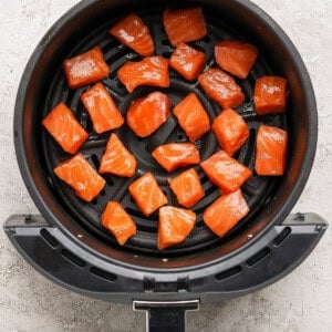 Raw salmon cubes are arranged in a single layer inside the basket of an air fryer on a light-colored surface.