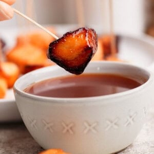 A hand holds a skewer with a glazed, roasted bite-sized food piece above a bowl of dipping sauce; more skewered pieces are seen in the background.