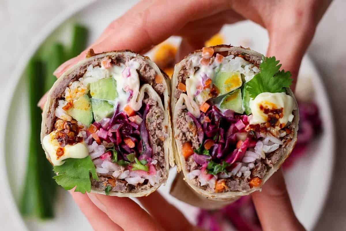 A close-up of hands holding a cut burrito filled with rice, ground meat, cucumber, egg, shredded vegetables, cilantro, and sauce, with a plate of food in the background.