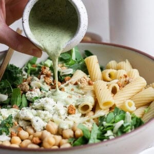 A hand pours creamy green dressing over a bowl of pasta salad with rigatoni, chickpeas, leafy greens, and crumbled toppings.