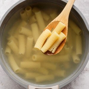 A pot of rigatoni pasta boiling in water, with a wooden spoon lifting a few pieces above the surface.