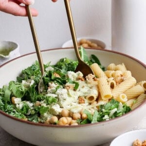 Hands using utensils to mix a salad with rigatoni pasta, greens, chickpeas, cheese, and dressing in a large bowl, with small bowls of ingredients nearby.