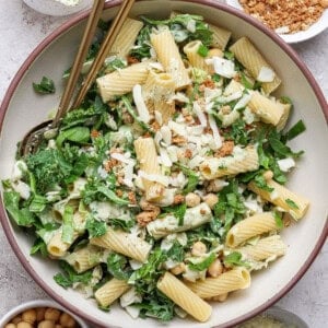 Large bowl of rigatoni pasta mixed with shredded greens, chickpeas, grated cheese, and creamy dressing, topped with breadcrumbs; serving utensils in the bowl.