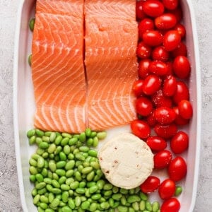 Raw salmon fillets, cherry tomatoes, edamame, and Boursin cheese assembled in a white casserole dish before baking