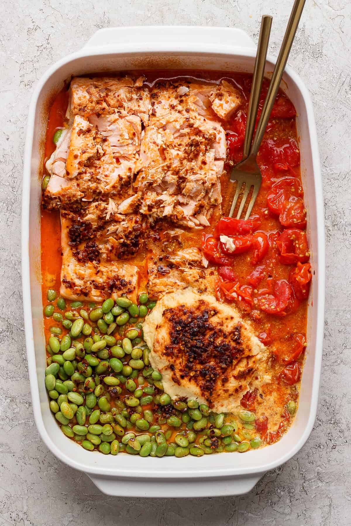 Baked salmon casserole with roasted tomatoes, edamame, and melted Boursin cheese with two gold forks in a white dish