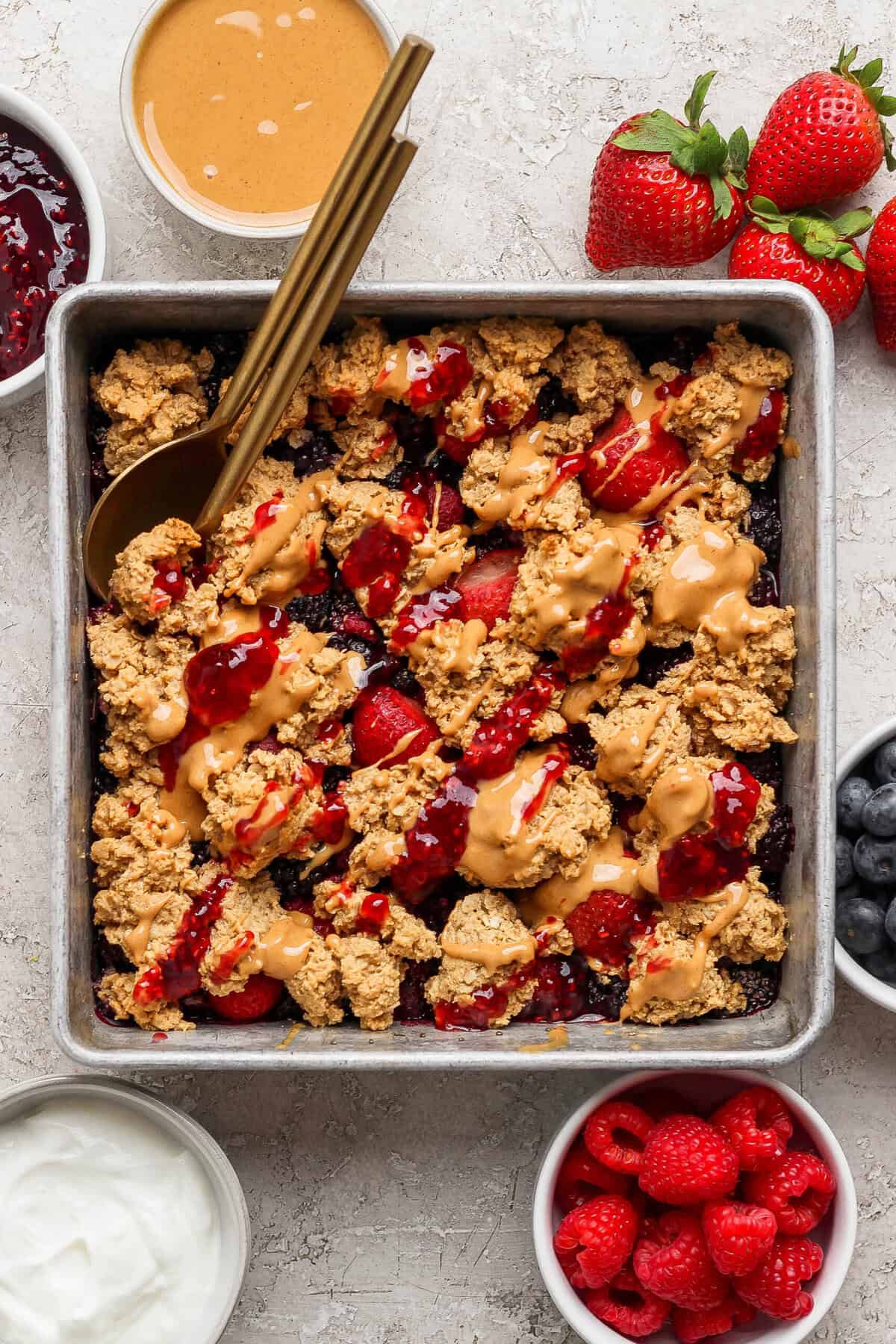 overhead view of protein berry crumble in a pan with peanut butter drizzle surrounded by bowls of jam yogurt and fresh berries