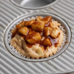 Unbaked muffin batter in a paper liner, topped with small cinnamon-glazed apple pieces, sits in a metal muffin pan.