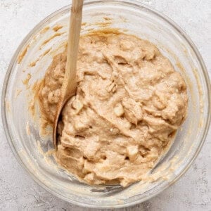 A glass bowl containing thick, chunky batter and a wooden spoon, set on a light-textured surface.
