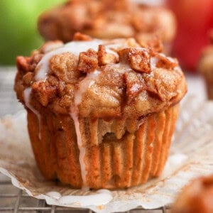 A muffin topped with small cinnamon apple pieces and white icing, sitting on an opened paper liner with a blurred background.