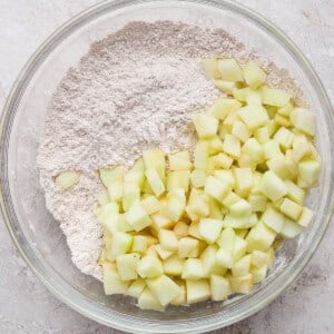 A glass bowl containing flour and diced green apples on a light textured surface.