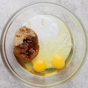 A glass bowl containing two eggs, brown sugar, honey, and a white liquid ingredient on a light textured surface.