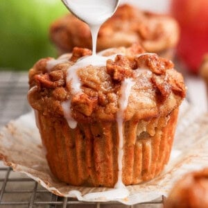A spoon drizzles white icing over a muffin topped with small pieces of fruit, set on a wire rack with an open paper liner.