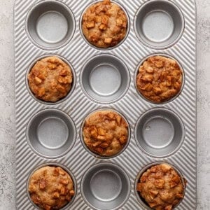A muffin tin containing six baked muffins with diced apple pieces, with three empty spaces in the tray, on a light textured surface.