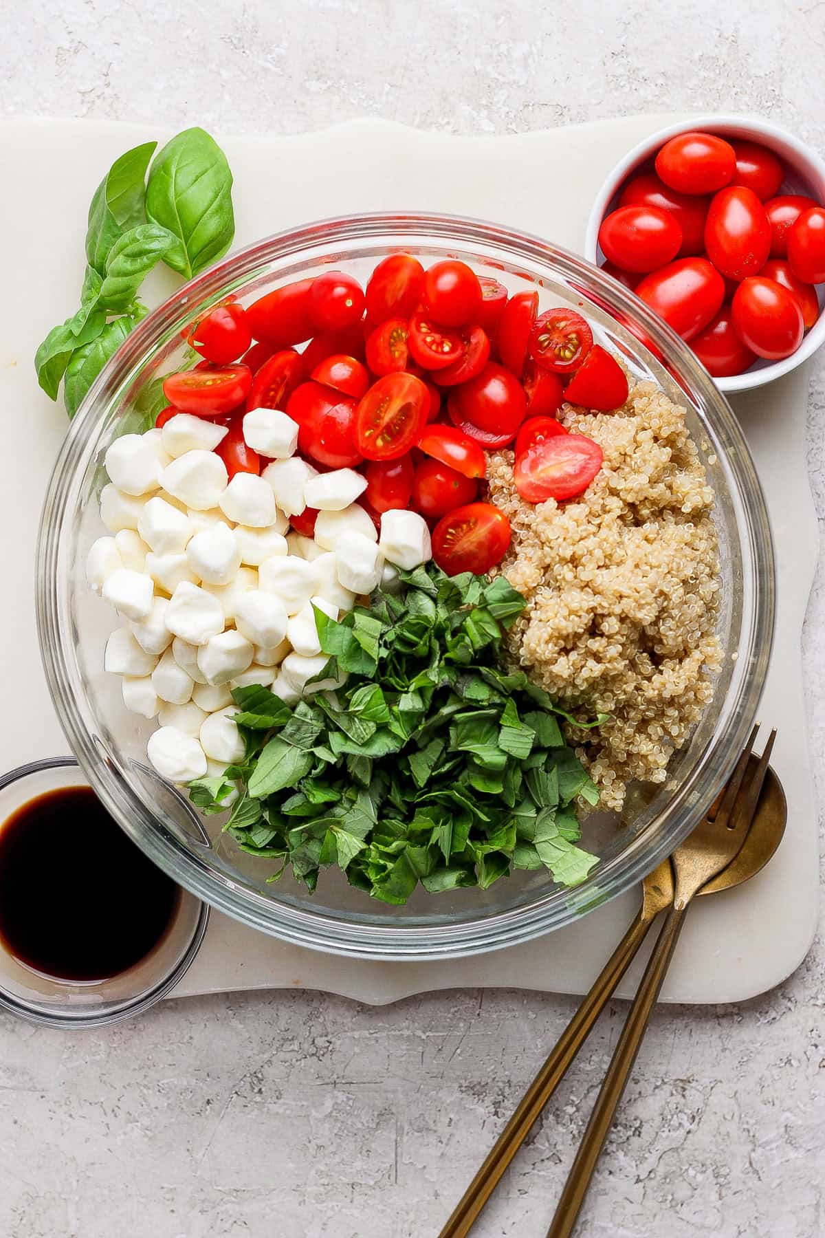 A glass bowl contains chopped basil, mozzarella balls, halved cherry tomatoes, and quinoa. Nearby are a small bowl of cherry tomatoes, basil leaves, a cup of dark liquid, and two gold utensils.