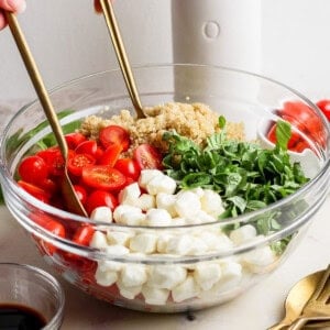 A person tosses a salad in a glass bowl with mozzarella balls, cherry tomatoes, quinoa, spinach, and basil. A bottle and small bowl of balsamic vinegar are nearby.