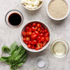 Bowls with cherry tomatoes, mozzarella balls, quinoa, balsamic vinegar, and oil, plus basil leaves and salt, arranged on a light surface.