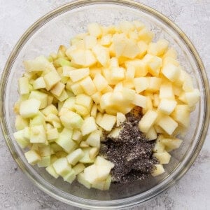 A glass bowl containing diced green and yellow apples with a pile of chia seeds on a light textured surface.