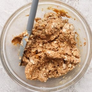 A glass bowl containing oatmeal cookie dough being mixed with a spatula on a light countertop.