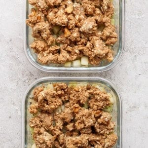 Two glass containers filled with unbaked fruit crisp, featuring a crumbly oat and nut topping over sliced fruit on a light stone surface.
