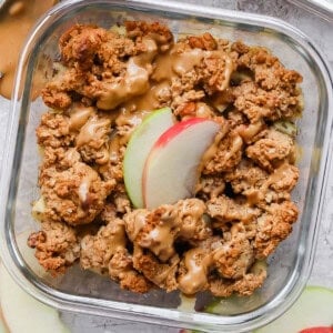 A glass container with apple slices, crumb topping, and a drizzle of light brown sauce. A spoon and extra apple slices are nearby.